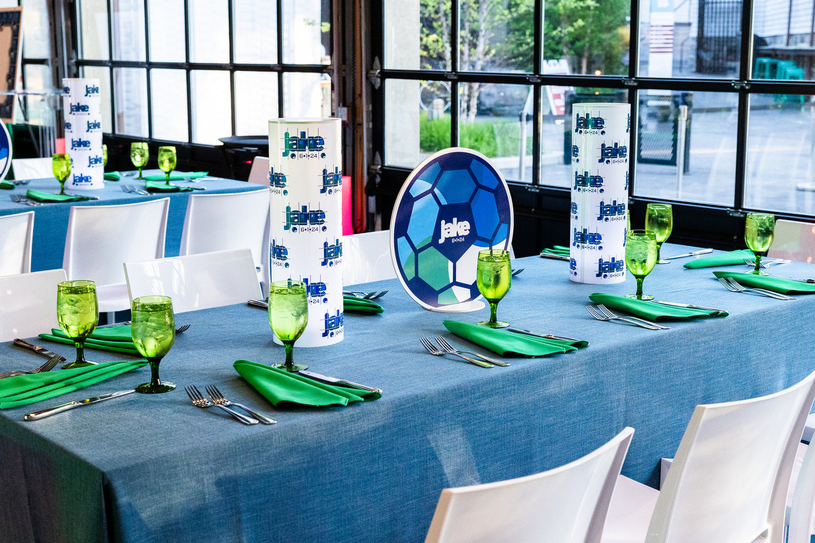 Table setting with green glassware, blue tablecloth, and branded napkins in a room with large windows.