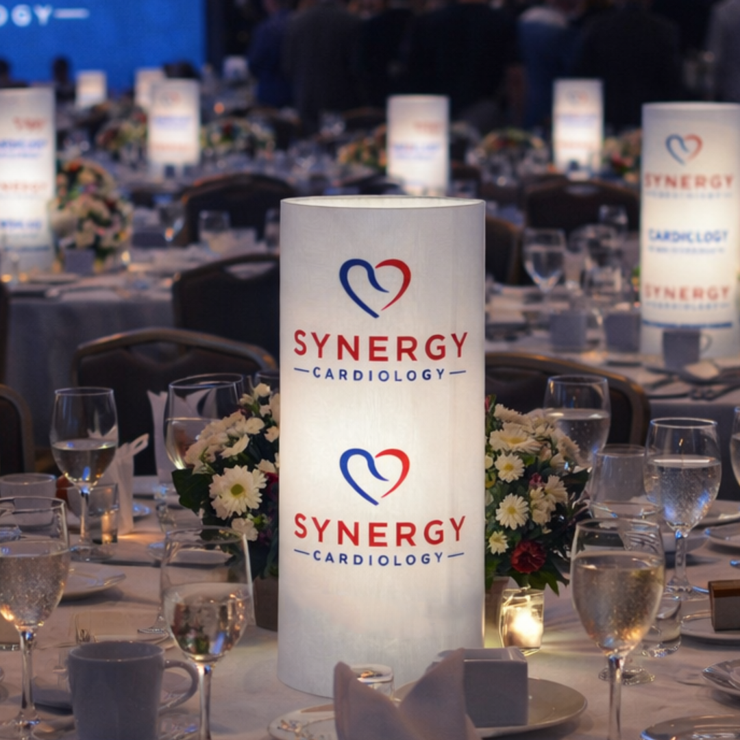 Synergy Cardiology branded table lamp on a dinner table with floral arrangements and wine glasses.