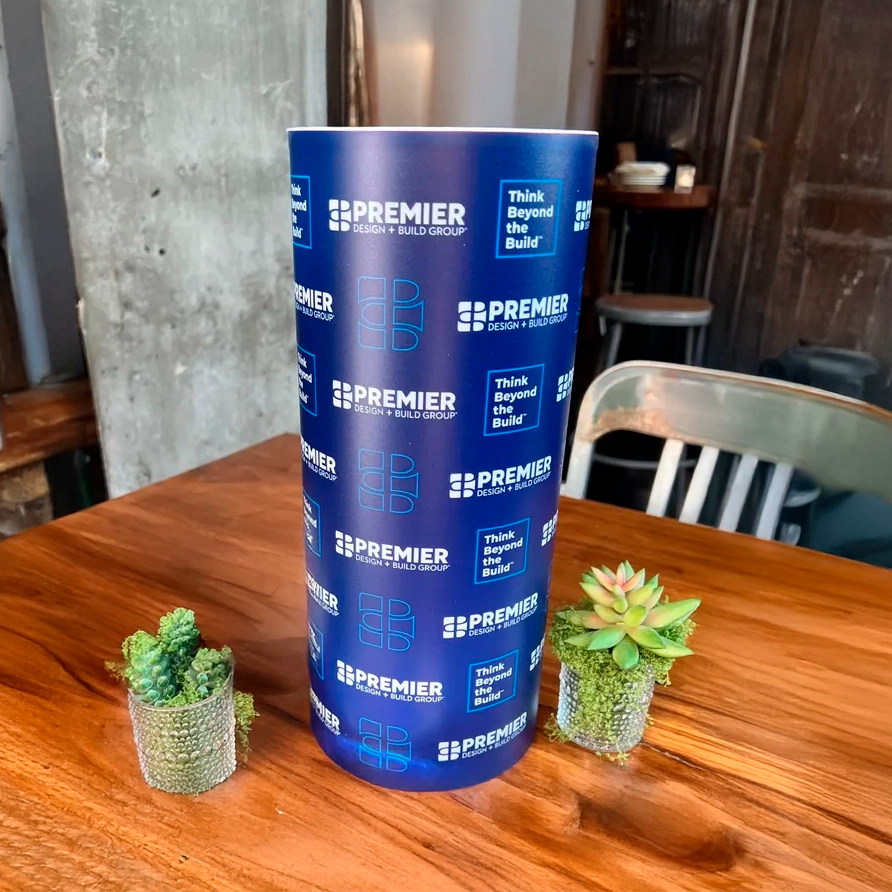 Blue cylindrical sign with 'Premier Design Build Group' branding on a wooden table with small potted plants.